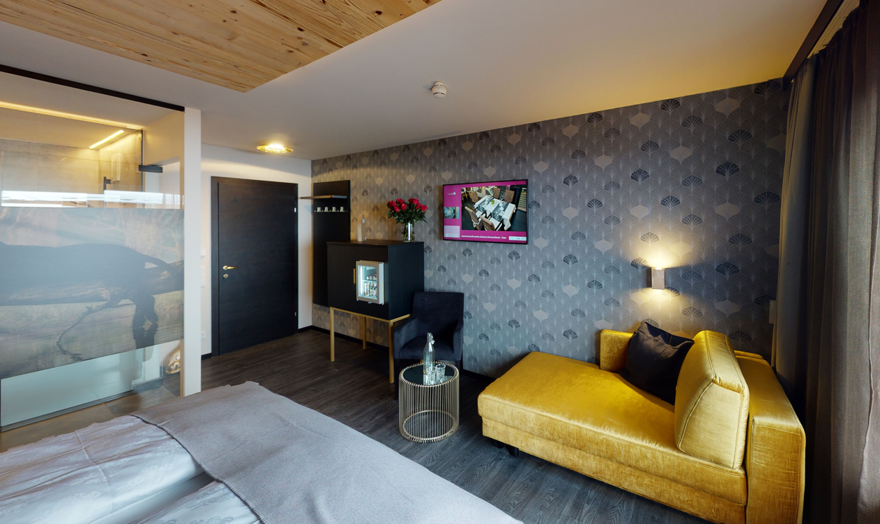 Et moderne hotelværelse med en mønstret accentvæg, en gul chaiselong, et væghængt tv, et lille glasbord og en minibar med blomster på toppen. Rummet har mørke gulve og loftspanel i træ.