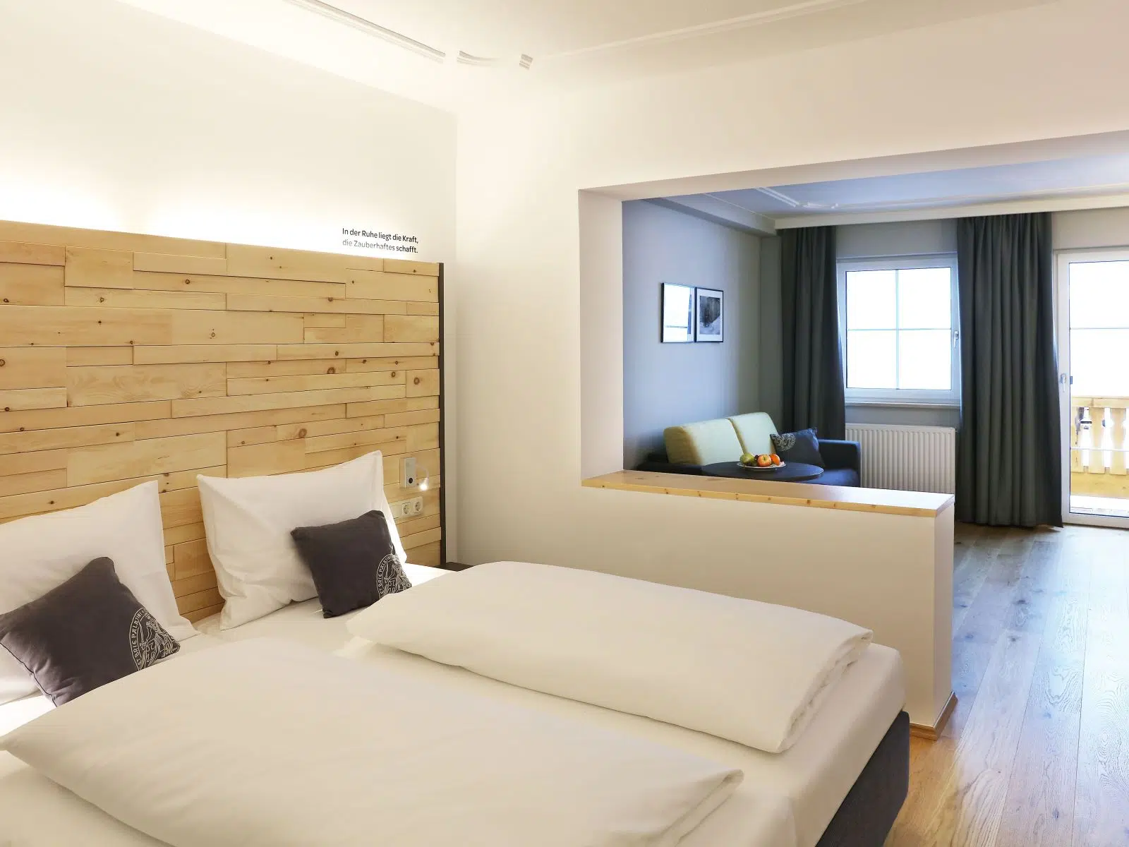 Et moderne hotelværelse med dobbeltseng, sengegavl af træ og hvidt sengetøj. Værelset har opholdsstue med sofa, store vinduespartier med gardiner og trægulv, hvilket skaber en lys og indbydende atmosfære.