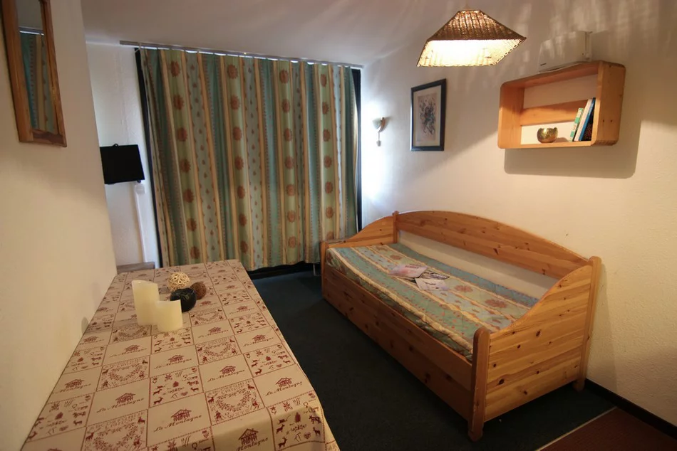 Et lille hyggeligt værelse med en daybed i træ, mønstrede gardiner, et bord med en printet dug, et par stearinlys og en væghylde med bøger. Blød belysning skaber en varm atmosfære.