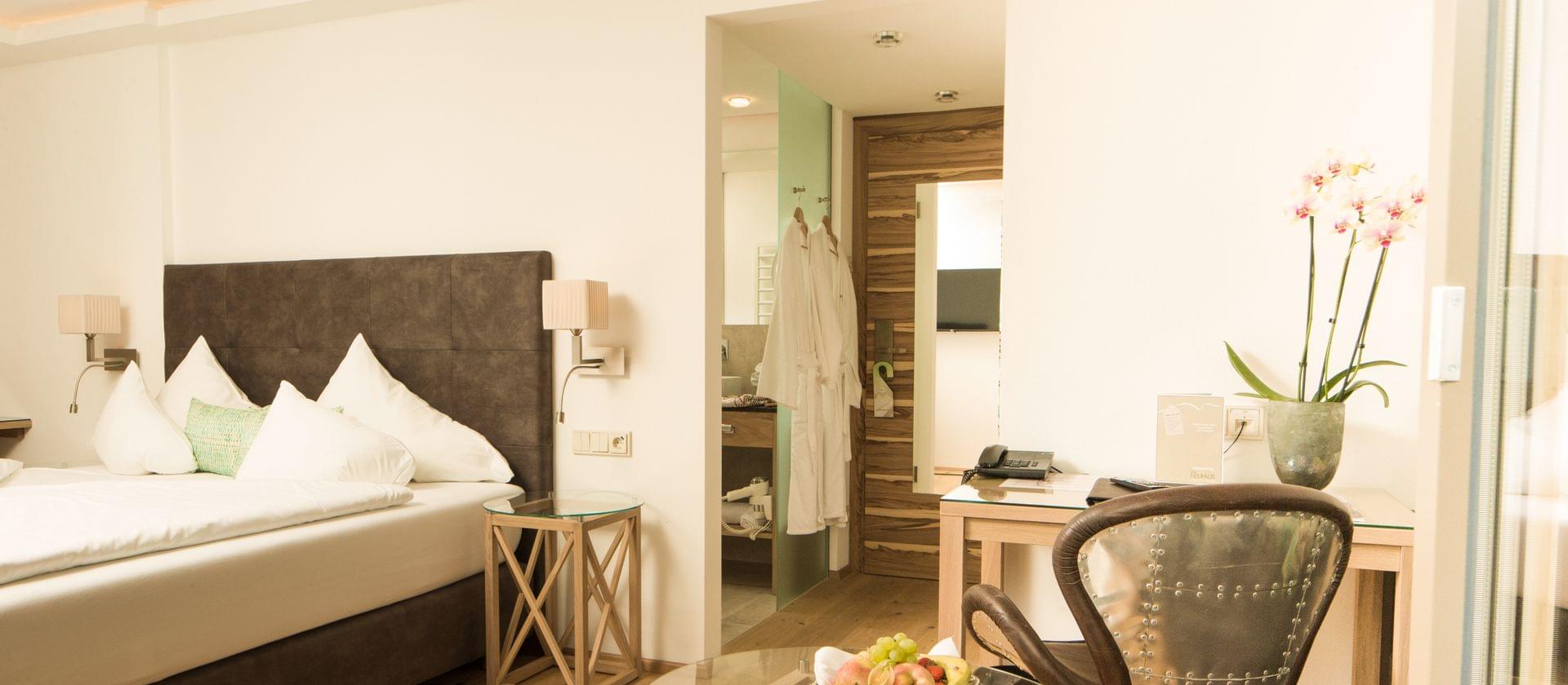 Lyst hotelværelse med pæn lavet dobbeltseng, bløde puder og moderne sidebord. Et skrivebord med en stol, telefon og vase med orkideer er i forgrunden. Badeværelse med morgenkåbe hænger i baggrunden.
