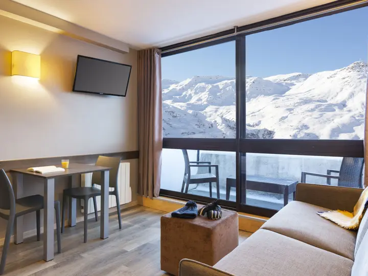 En hyggelig stue med bord, stole og sofa ud mod et stort vindue. Udsigten viser snedækkede bjerge under en klar blå himmel. Et vægmonteret tv er til venstre, og lyset strømmer ind og oplyser rummet.