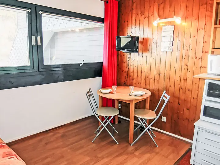 En hyggelig spiseplads har et lille træbord med tallerkener og glas, omgivet af to klapstole. Rummet er indrettet med træpaneler, røde gardiner og et lille væghængt tv. En mikrobølgeovn sidder på nærliggende hylder.