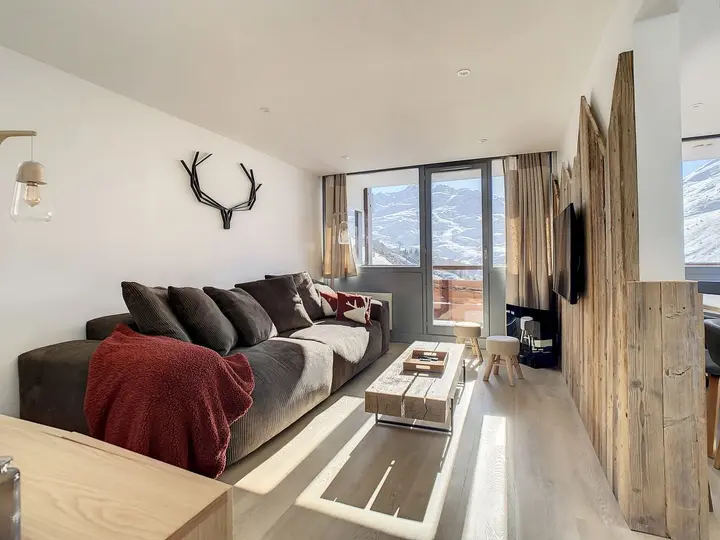En hyggelig stue med en stor brun sofa, rødt tæppe, sofabord i træ og gevir vægindretning. Værelset har naturlige træaccenter og udsigt over bjergene gennem et stort vindue. Lyse trægulve fremhæver det lyse, luftige rum.