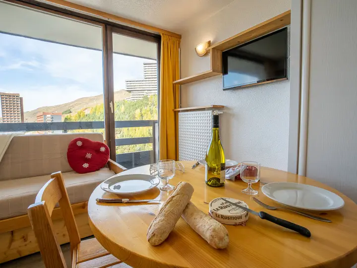 En hyggelig spiseplads med et rundt træbord til to. Den byder på tallerkener, glas, en flaske vin og baguettes. En sofa med en hjerteformet pude er ved et vindue, der afslører en naturskøn udsigt over bjergene under en klar himmel.