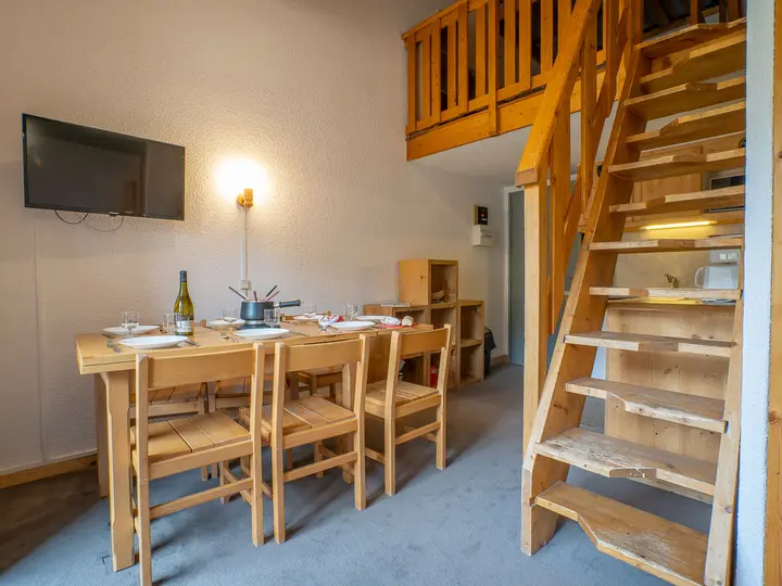 En hyggelig spiseplads med et træbord til fire, med en fonduegryde og vinflaske. En trappe fører til en hems ovenpå, og et væghængt tv er i nærheden. Rummet er varmt oplyst med en væglampe.