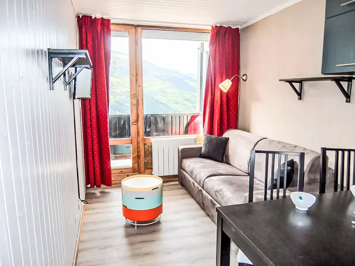En hyggelig stue med en grå sofa, røde gardiner og et stort vindue med en naturskøn udsigt over bjergene. Et lille rundt bord og to sorte stole er tæt på et mørkt spisebord. Vægmonteret tv og hylder giver ekstra opbevaring.