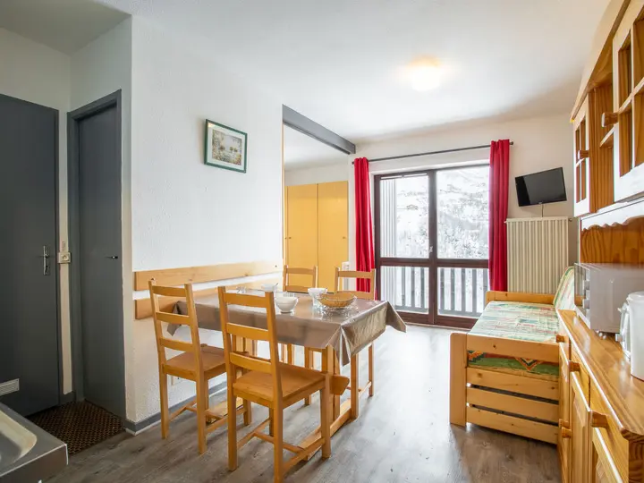 En hyggelig opholds- og spisestue med et træbord og -stole, en polstret bænk, en skænk med køkkenting og et vindue med røde gardiner med udsigt over sne. Rummet er minimalistisk indrettet med et billede på væggen.