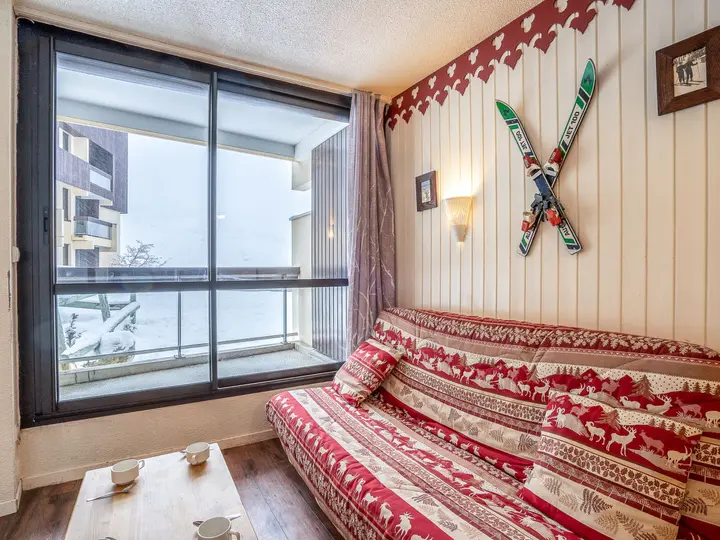Hyggeligt værelse med indretning med ski-tema, med en rødmønstret sovesofa op ad en trævæg dekoreret med krydsede ski og små indrammede billeder. En stor glasskydedør tilbyder en sneklædt udsigt udenfor. Fire kopper hviler på et sofabord af træ.