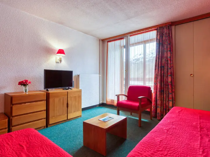 Et hyggeligt hotelværelse med to røddækkede senge og en tilhørende lænestol. Et fladskærms-tv sidder på en trækommode. Et lille sofabord i træ rummer et hæfte. Et vindue med blomstergardiner afslører en sneklædt bjergudsigt udenfor.