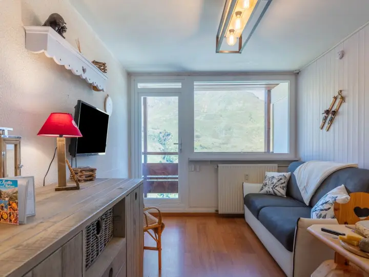 Hyggelig stue med sofa, monteret tv og træskab. En rød lampe sidder på skabet, og dekorative ski hænger på væggen. Et stort vindue viser bjergudsigt, og en balkondør tilføjer naturligt lys.