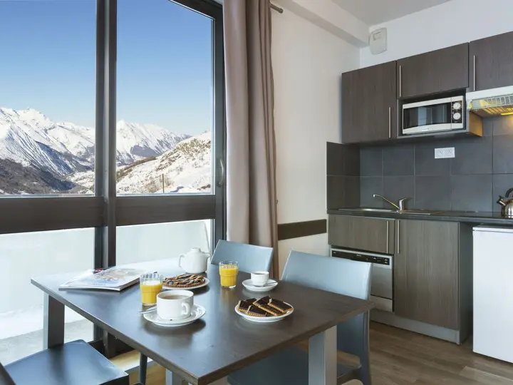 Moderne køkken med et spisebord til morgenmad, med appelsinjuice, kaffe og kageskiver. Store vinduer giver en malerisk udsigt over snedækkede bjerge under en klar blå himmel.