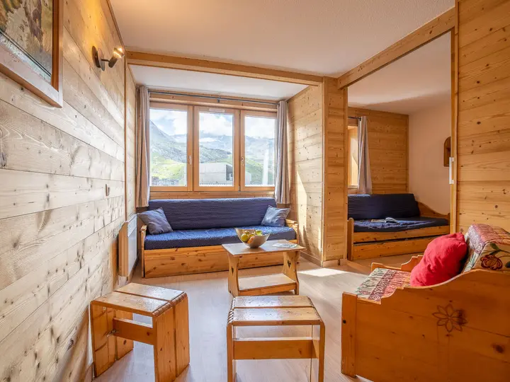 En hyggelig træstue med store vinduer, to blå polstrede bænke, en træstol med farverige puder, et lille bord og stærkt naturligt lys, der kommer ind og afslører en bjergudsigt udenfor.