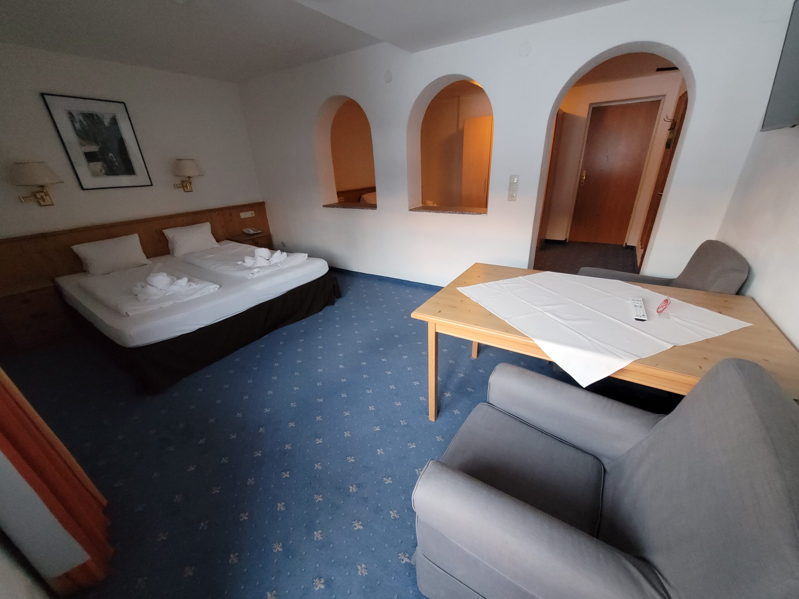 Et hotelværelse med en dobbeltseng, to hvide morgenkåber, et træbord med en hvid dug, en grå lænestol, blåt tæppe og buede åbninger i væggen, der fører til et andet område.