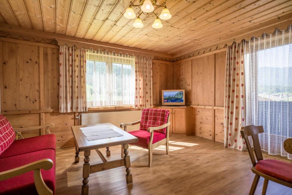 En hyggelig træstue med røde polstrede stole, et træbord, TV på et skab, store vinduer med gardiner og sollys, der strømmer ind og skaber en varm og indbydende atmosfære.