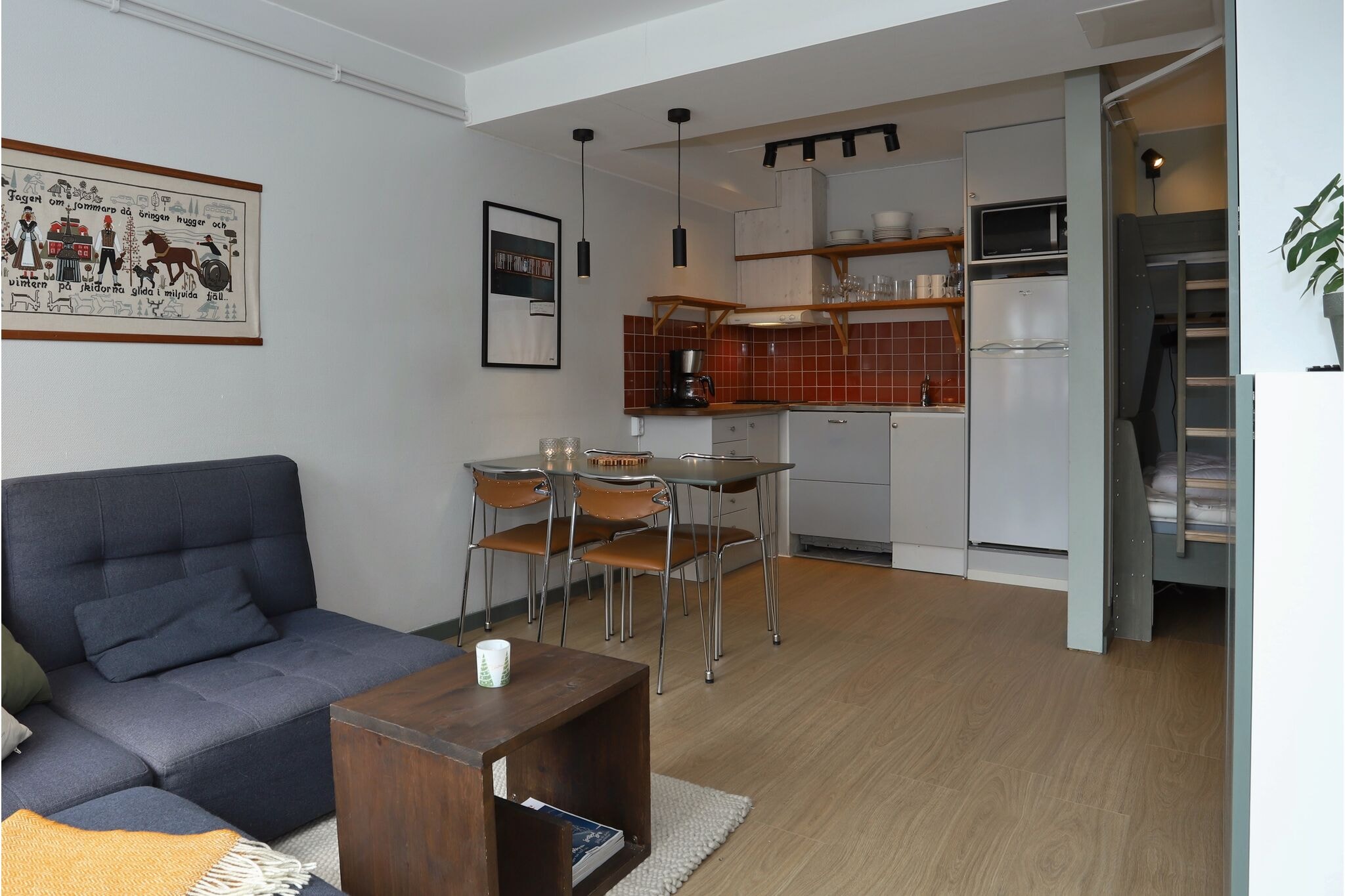 Moderne studiolejlighed med en grå sofa, et lille træbord, spiseplads til fire personer, åbent køkken med rød flisebelægning, hvide apparater og en køjeseng, der delvist er synlig til højre. Minimalistisk indretning og trægulv.