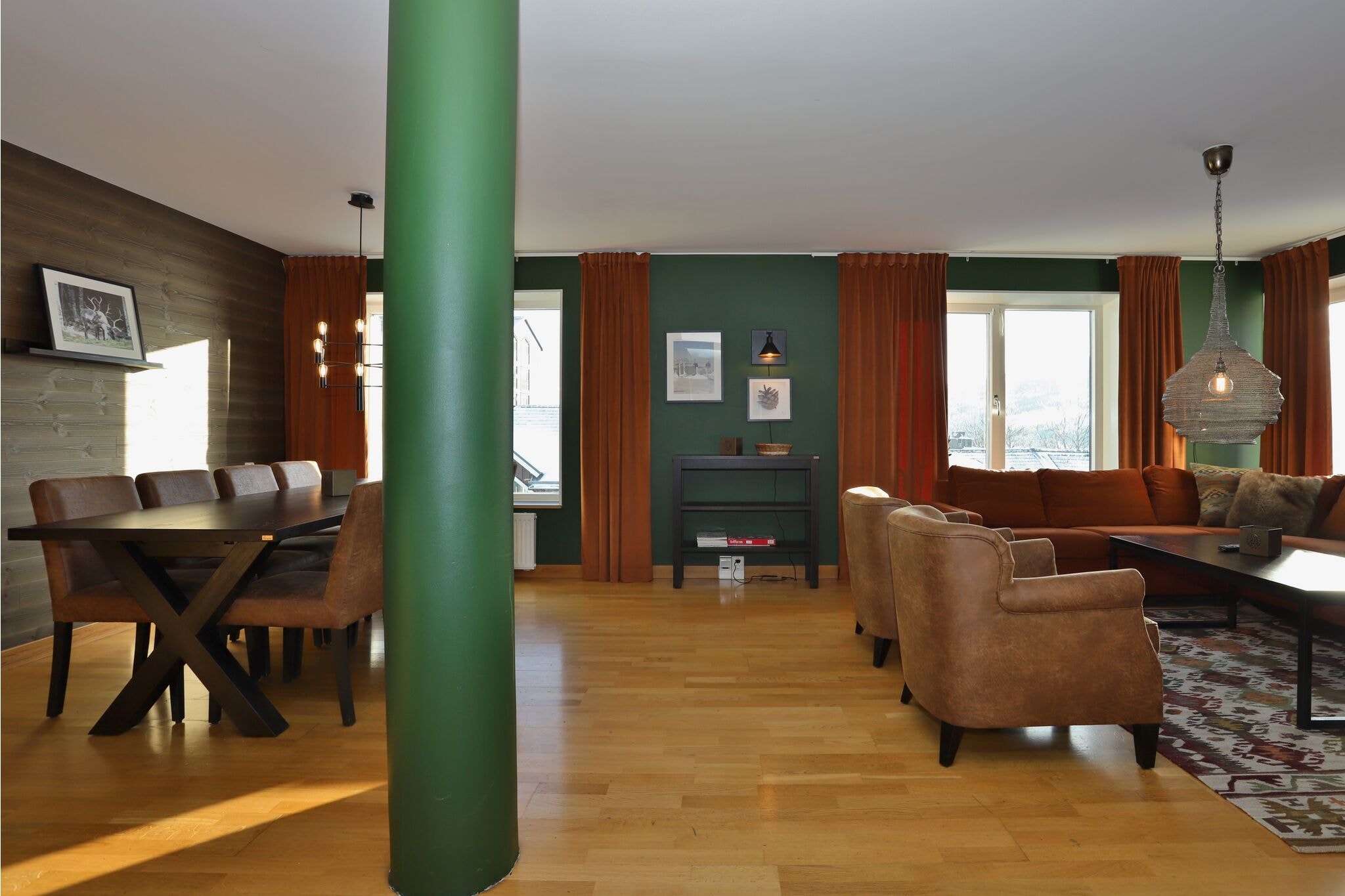 Moderne opholds- og spisestue med trægulve, grøn accentvæg, orange gardiner, en grøn søjle, brune lænestole, en sektionssofa, vægkunst og et spisebord med polstrede stole. Store vinduer giver naturligt lys.