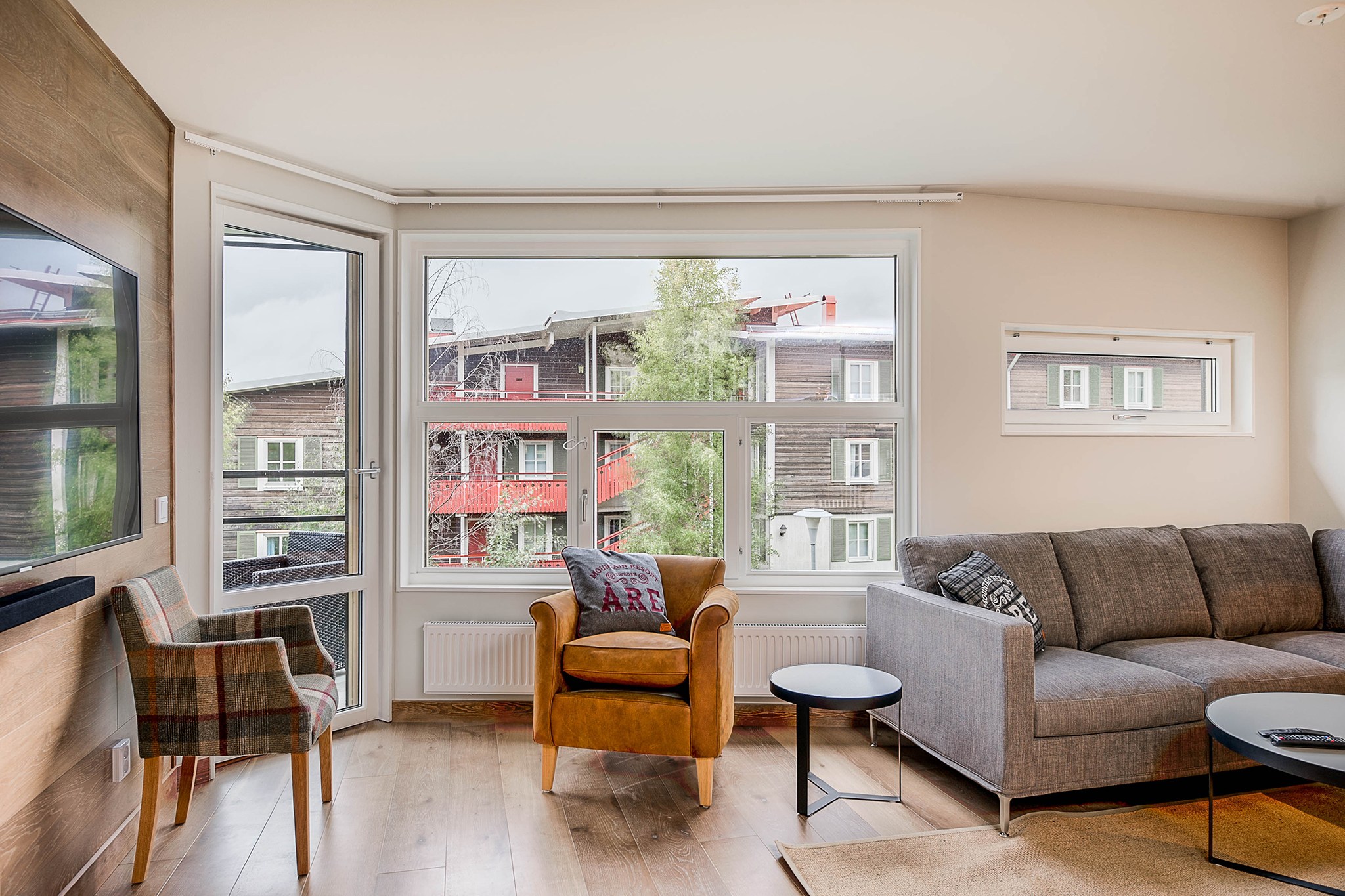 En lys stue med store vinduer, en grå sofa, en beige lænestol, en ternet stol, små borde og et vægmonteret tv. Udenfor er der synlige trælejlighedsbygninger og træer.
