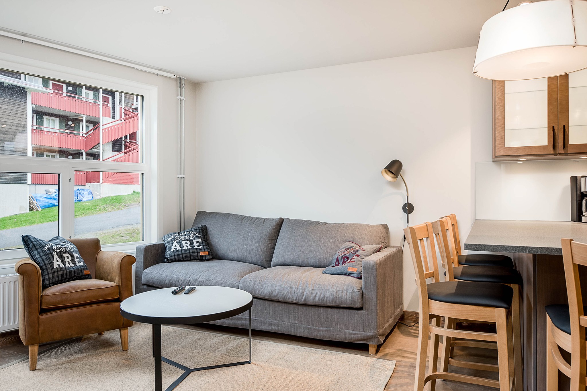 En moderne stue med en grå sofa, en lænestol, et rundt sofabord og fire barstole i træ ved en disk. En gulvlampe står ved siden af sofaen, og et stort vindue viser en lejlighedsbygning udenfor.