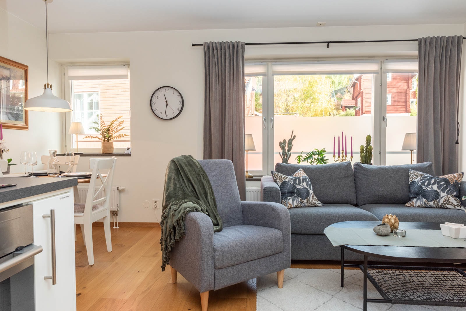 En lys, moderne stue med en grå lænestol og sofa med mønstrede puder, et sofabord, store vinduer med gardiner og en spiseplads med bord og stole. Planter og indretning tilføjer hyggelige detaljer.