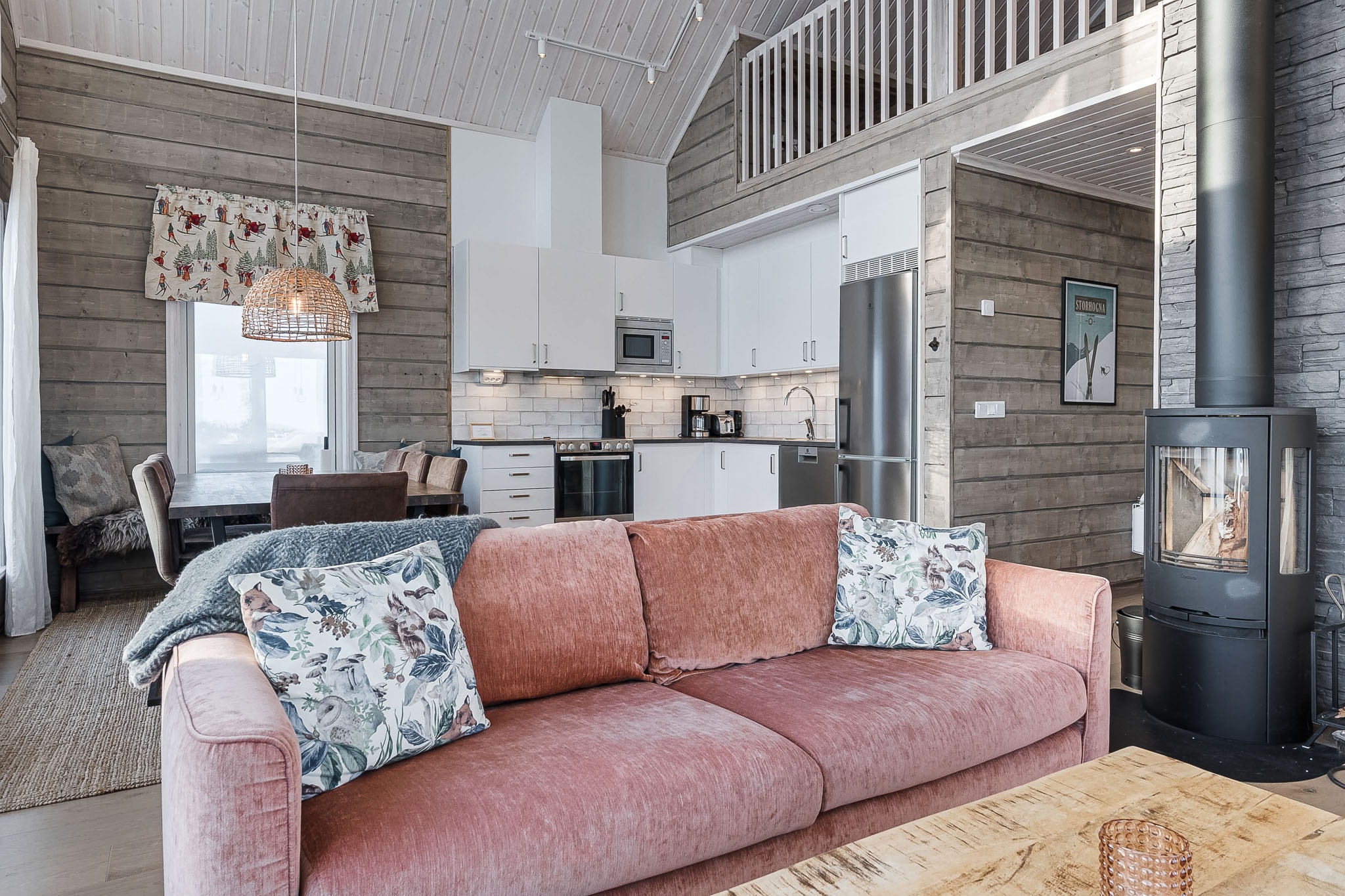 En hyggelig stue med en lyserød sofa, blomstrede puder, en brændeovn og et åbent køkken med hvide skabe. Trævægge og -lofter skaber en varm, rustik atmosfære. Spiseområdet ses i baggrunden.