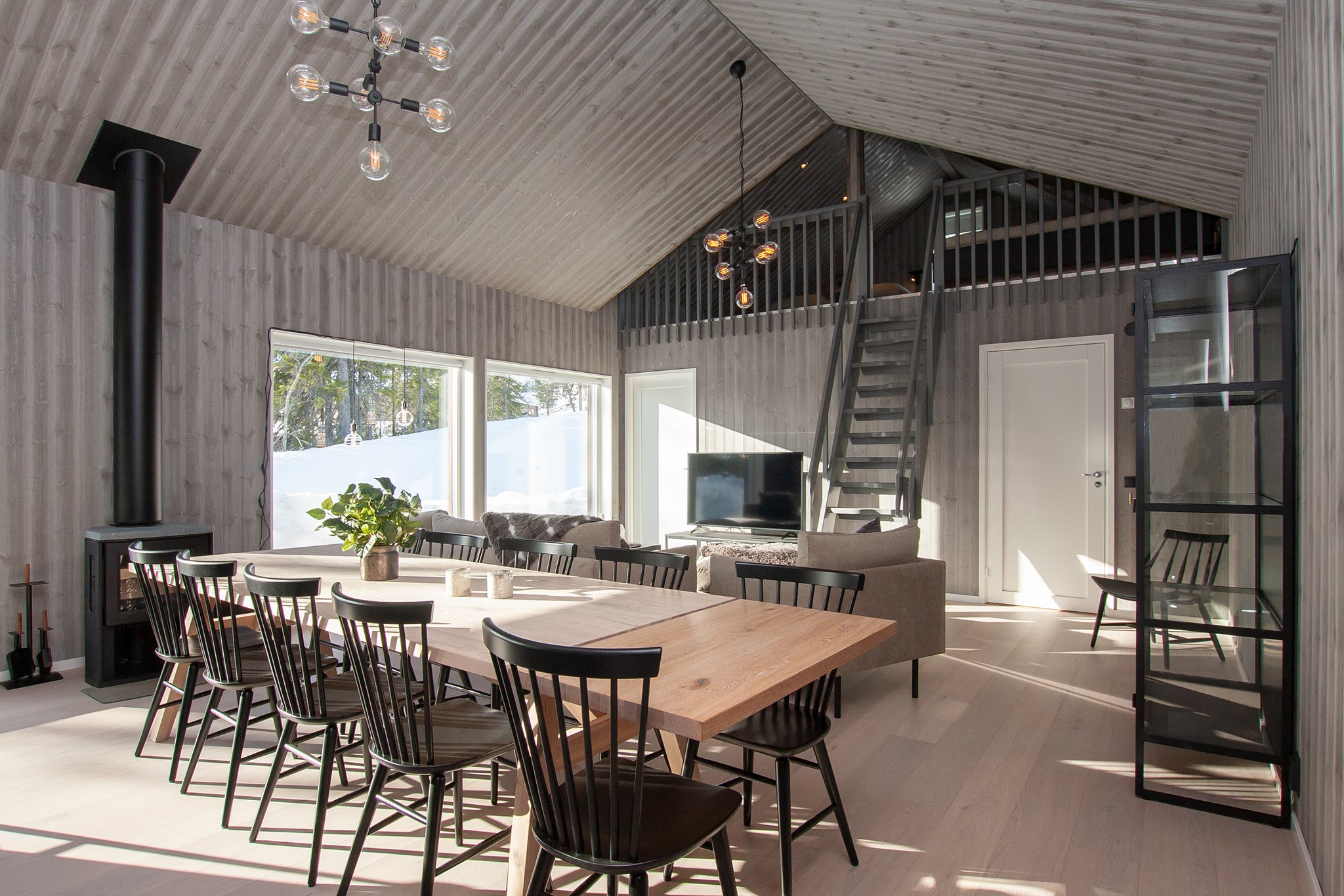 Moderne, solbeskinnet åben stue og spisestue med træpaneler på vægge og i loftet, store vinduer, sorte stole omkring et træbord, grå sofa, trappe til hems og sort brændeovn i hjørnet.