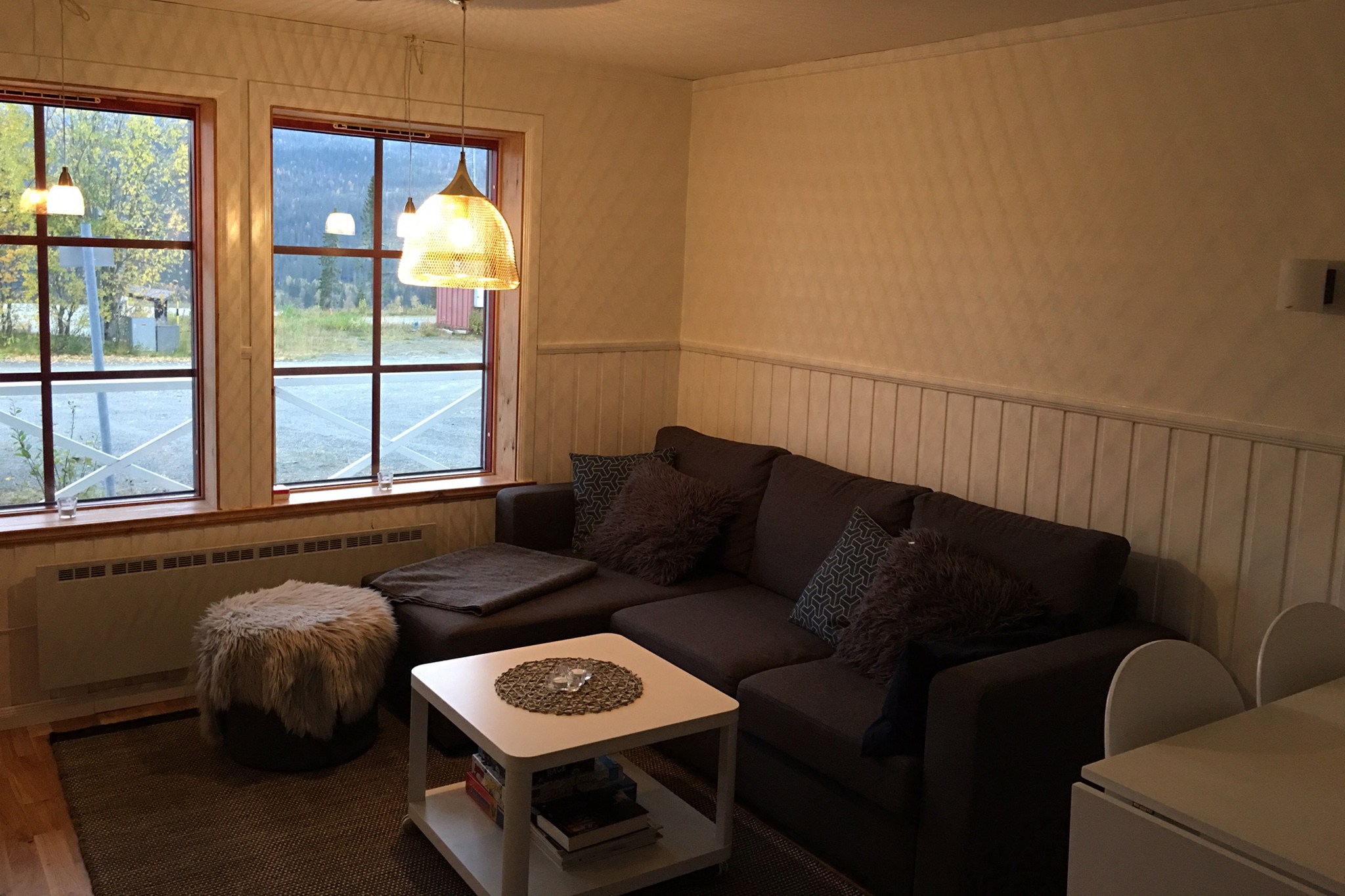 En hyggelig stue med en mørk sofa, bløde puder, en lille puf, et hvidt sofabord og to store vinduer, der lukker dagslyset ind. En pendellampe hænger fra loftet, og der er udsigt til træer og en vej udenfor.