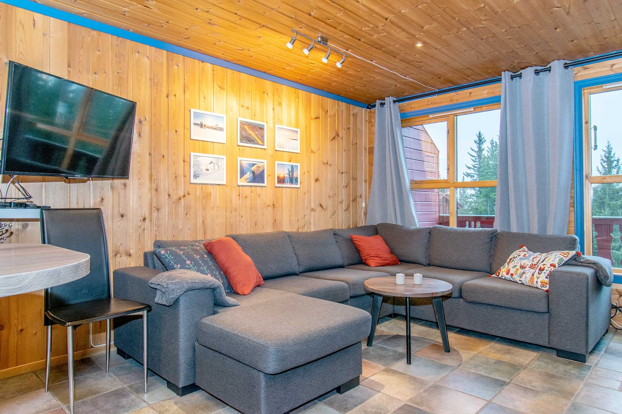 Hyggelig stue med trævægge og -loft, grå sofa med farverige puder, rundt sofabord, vægmonteret TV, indrammede fotos på væggen, store vinduer med grå gardiner og flisebelagt gulv.