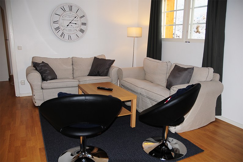 En hyggelig stue med to beige sofaer, et sofabord i træ, to sorte moderne stole, en gulvlampe, et stort vægur og et vindue med mørke gardiner. En fjernbetjening til tv'et ligger på bordet.