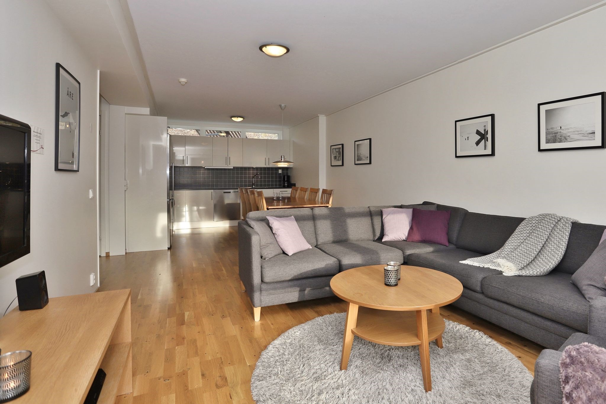 En moderne stue med en grå sofa, lilla og grå hynder, et rundt sofabord i træ på et rundt tæppe og indrammede fotos på væggen. I baggrunden er der en spiseplads og et køkken.