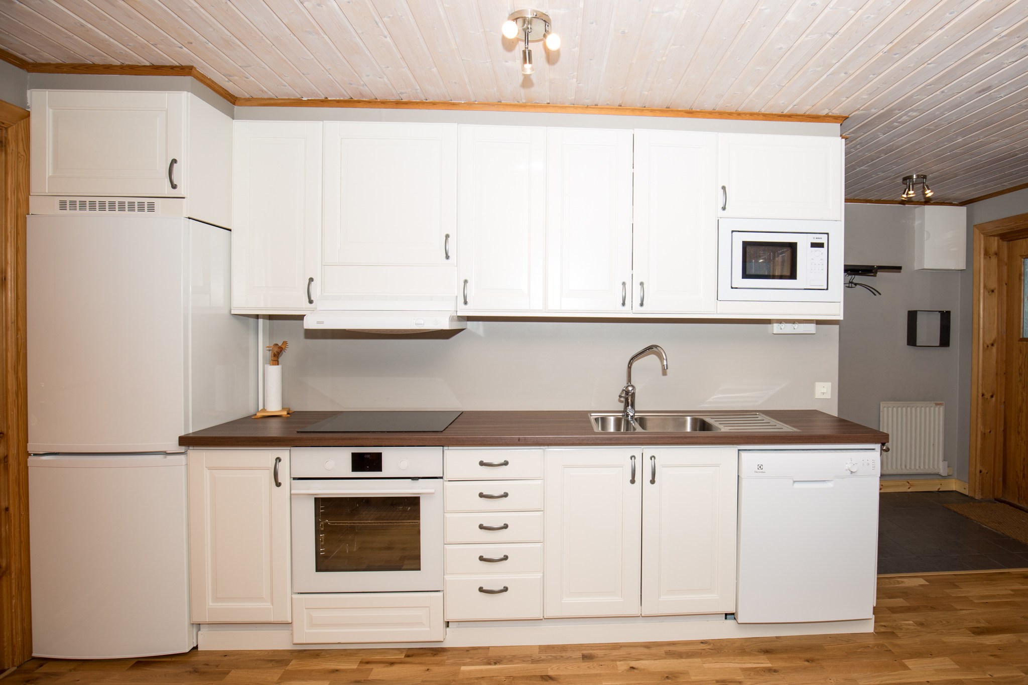 Moderne køkken med hvide skabe, køleskab, ovn, komfur, opvaskemaskine, mikrobølgeovn og vask. Bordpladen er brun, og loft og gulv er af træ. Rummet fremstår lyst og rent.