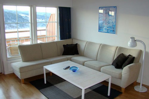 En moderne stue med en beige hjørnesofa, sorte og brune hynder, et hvidt sofabord med fjernbetjening og en blå tallerken, en gulvlampe, vægkunst og store vinduer, der viser et snedækket landskab udenfor.
