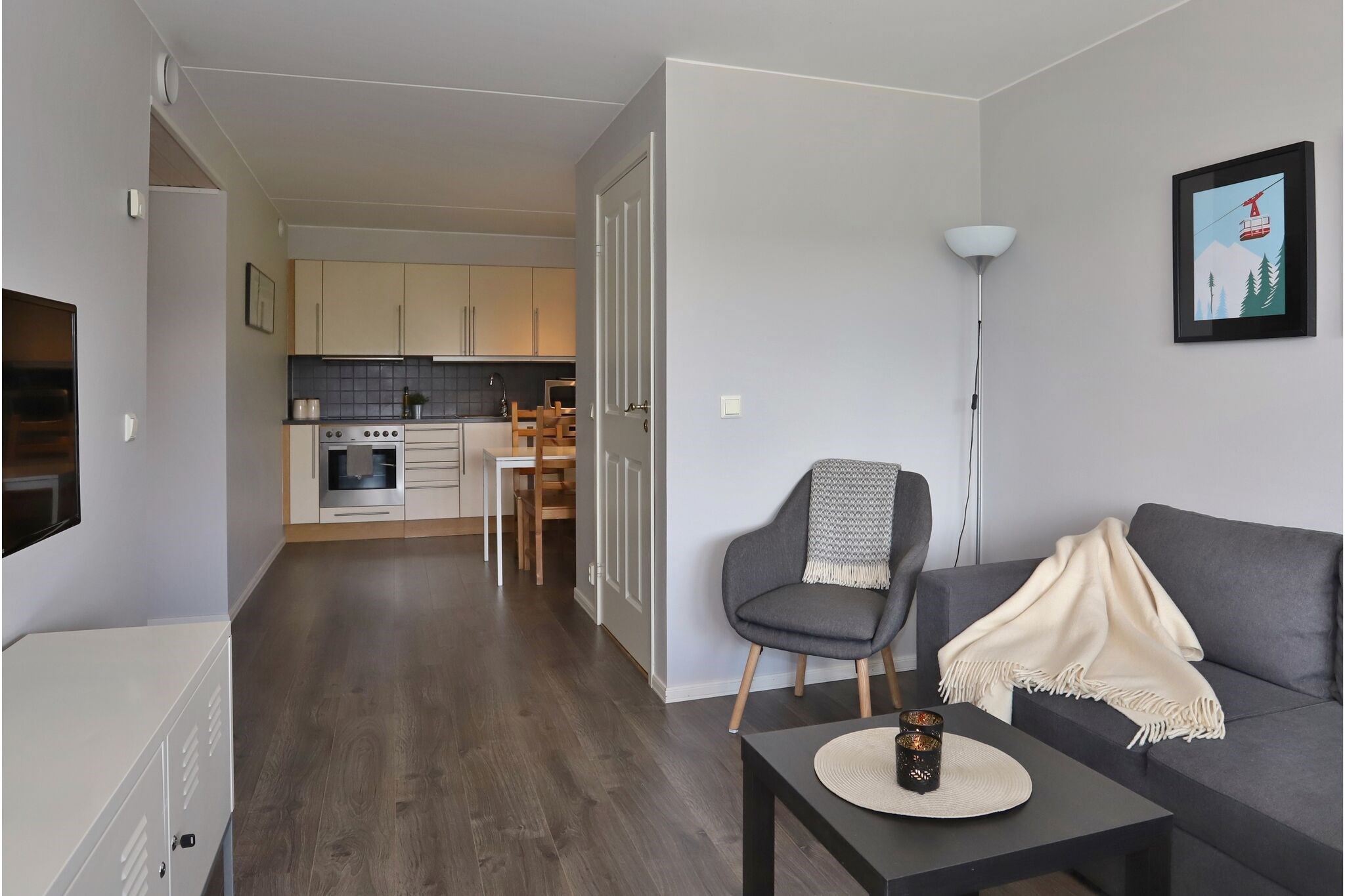 En moderne, minimalistisk stue med grå vægge, en grå sofa, lænestol, gulvlampe og vægkunst. Rummet åbner op til et lille køkken og spiseplads med lyse skabe og et spisebord.
