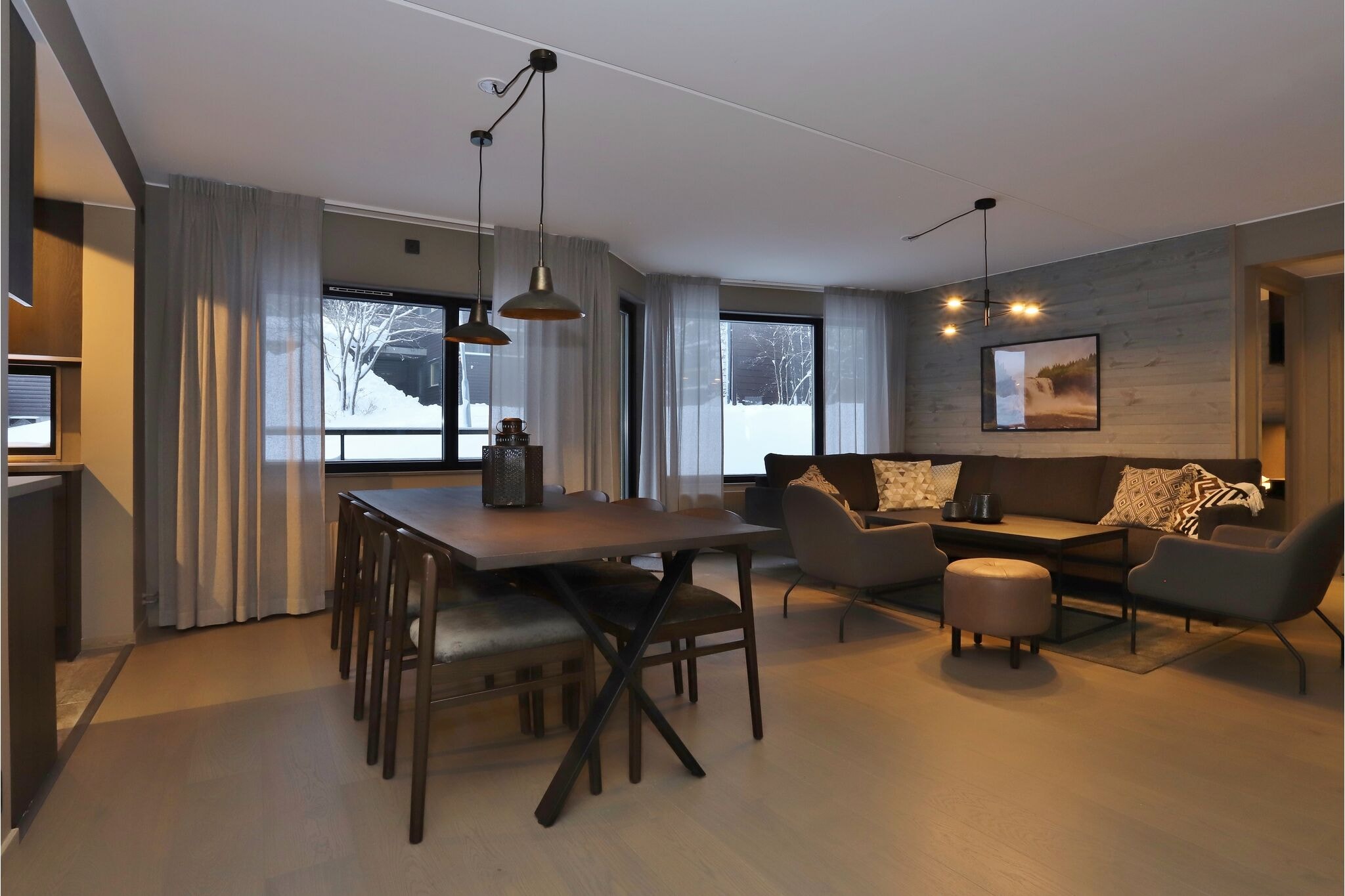 Moderne åben stue og spisestue med træbord, sorte stole, hyggelig sofa, lænestole, sofabord, blød belysning, store vinduer med gardiner og et synligt snelandskab udenfor.