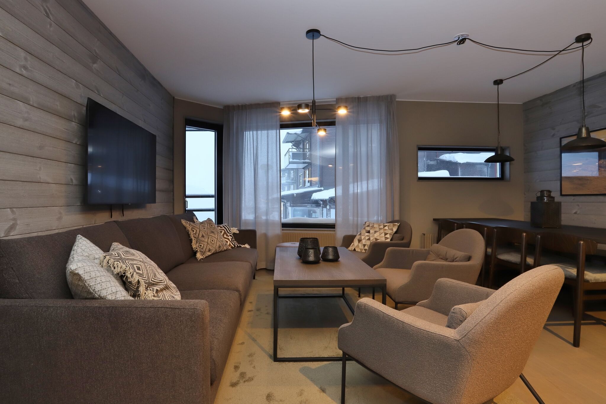 En moderne stue med grå sofaer, lænestole og et sofabord i træ. Store vinduer lukker naturligt lys ind og viser et snedækket landskab udenfor. Neutrale toner og minimalistisk indretning skaber en hyggelig atmosfære.