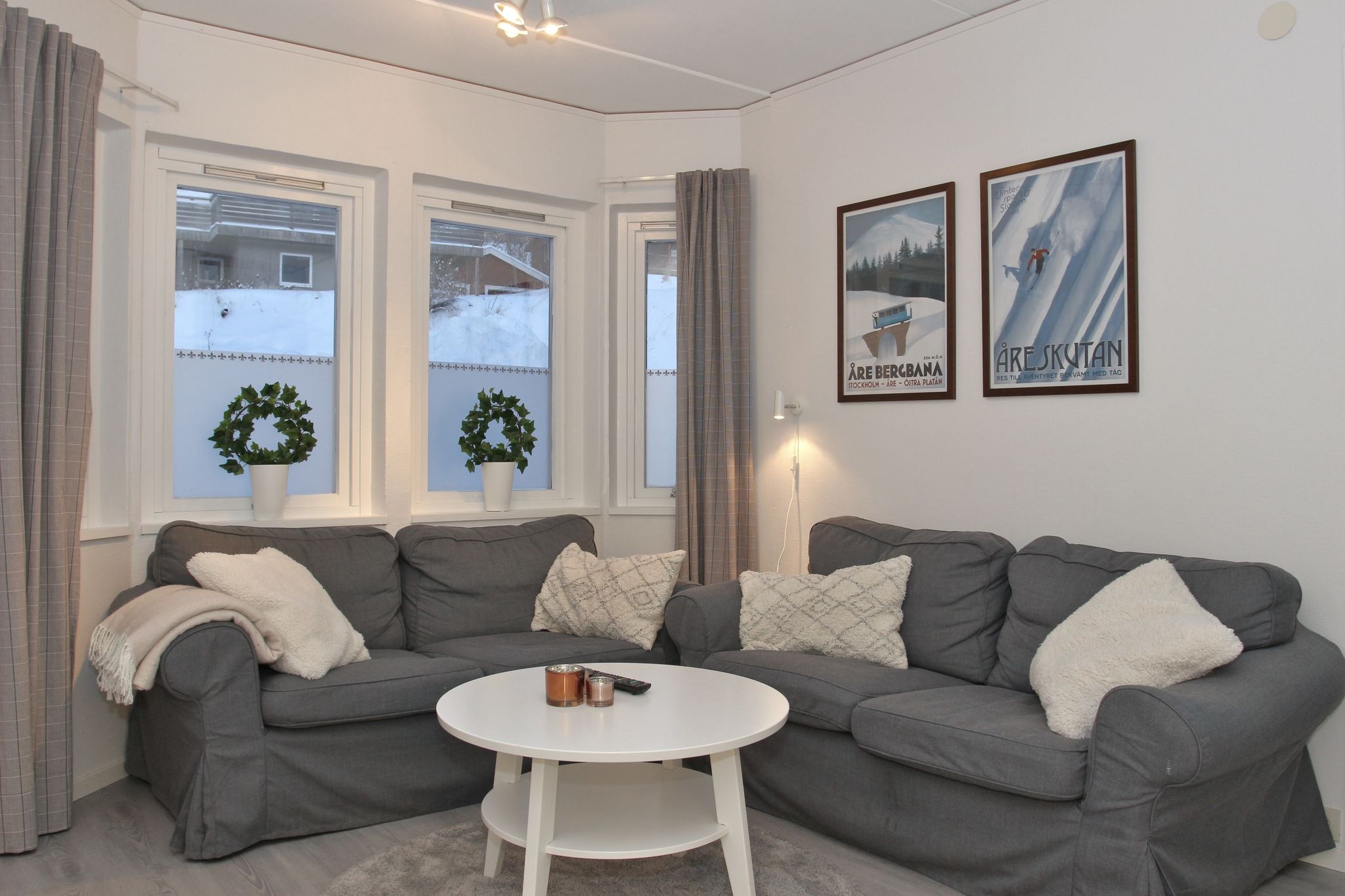En hyggelig stue med to grå sofaer, hvide hynder, et rundt hvidt sofabord, to potteplanter i vindueskarmen og vintage skiplakater på væggen. Sneen er synlig uden for vinduerne.