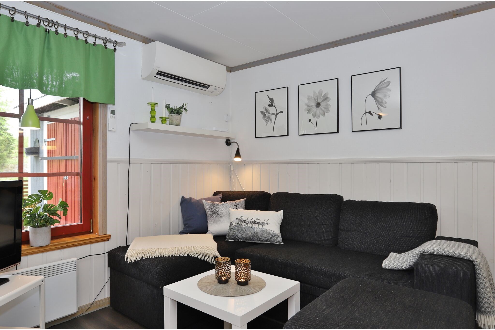 En hyggelig stue med en mørk hjørnesofa, et hvidt sofabord, stearinlys, grønne gardiner, potteplanter og tre sort-hvide blomstertryk på væggen over sofaen.