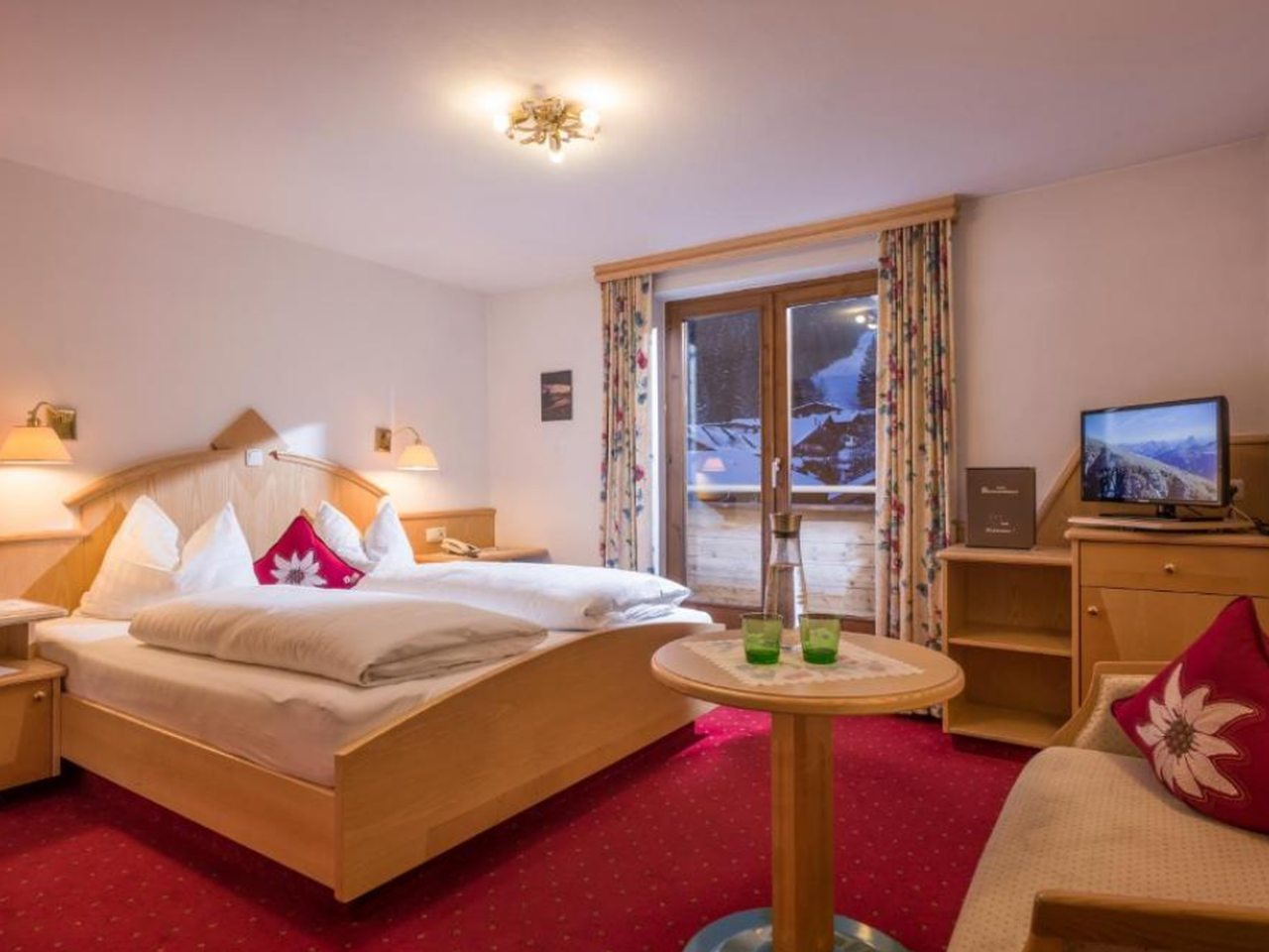 Et hyggeligt hotelværelse med dobbeltseng, rødt tæppe, træmøbler, et lille rundt bord med drinks, en sofa og en balkondør med udsigt over sneklædte bjerge udenfor. Til højre er der et tv og et skrivebord.