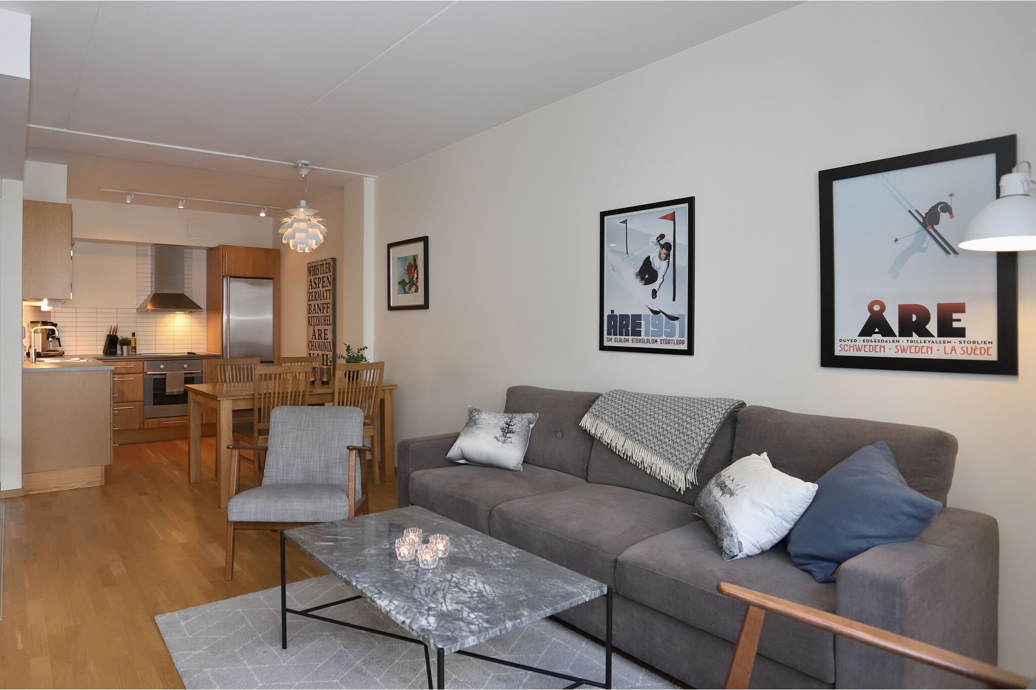 Moderne stue med en grå sofa, lænestol og marmor sofabord på et lyst tæppe. Spiseplads og køkken med træmøbler i baggrunden. Væggene er dekoreret med indrammet kunst og fotos. Varm, hyggelig belysning.