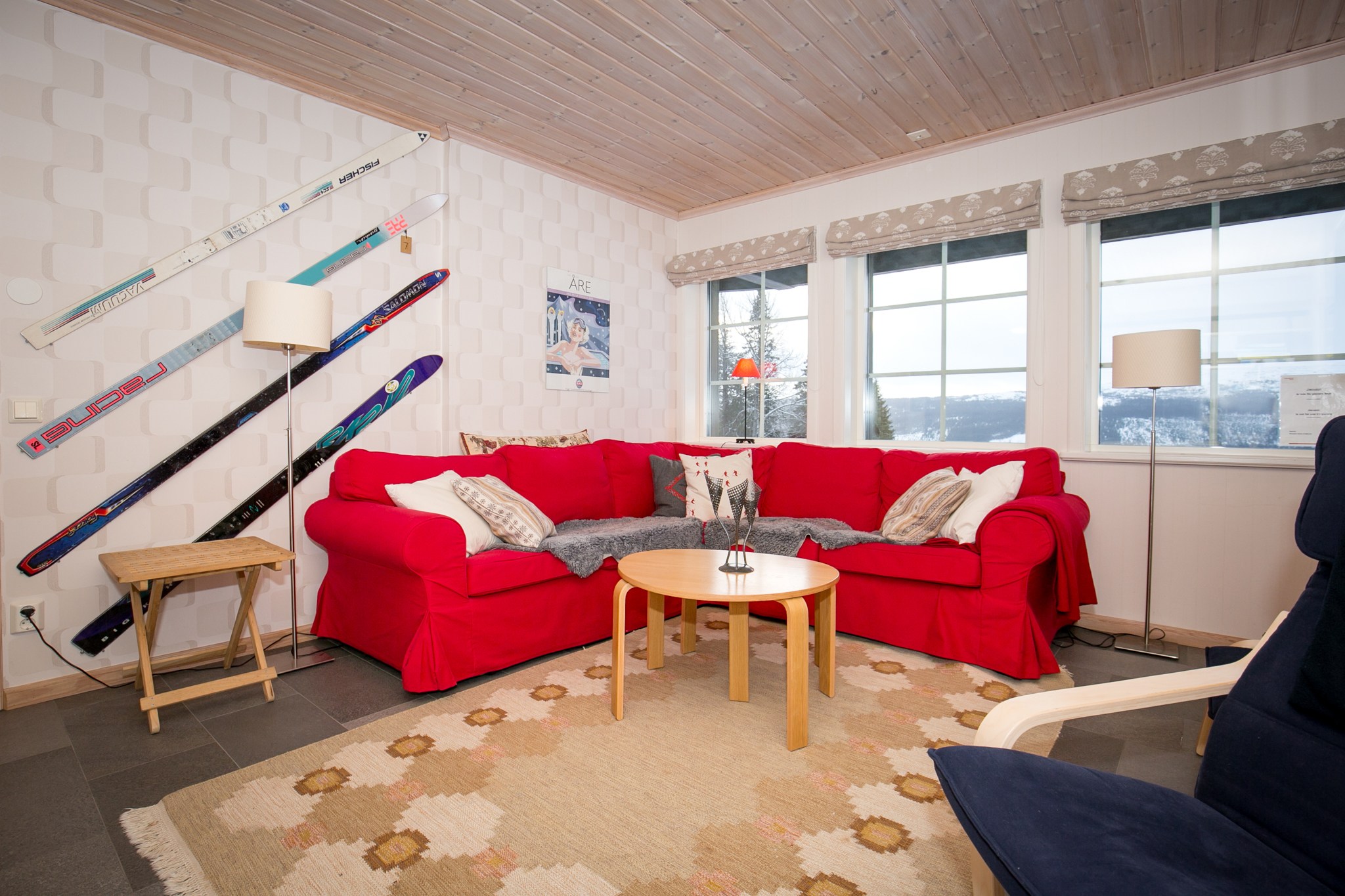 En hyggelig stue med to røde sofaer omkring et rundt sofabord i træ, et mønstret tæppe og vintageski monteret på væggen. Store vinduer lukker naturligt lys ind og tilbyder udsigt udad.