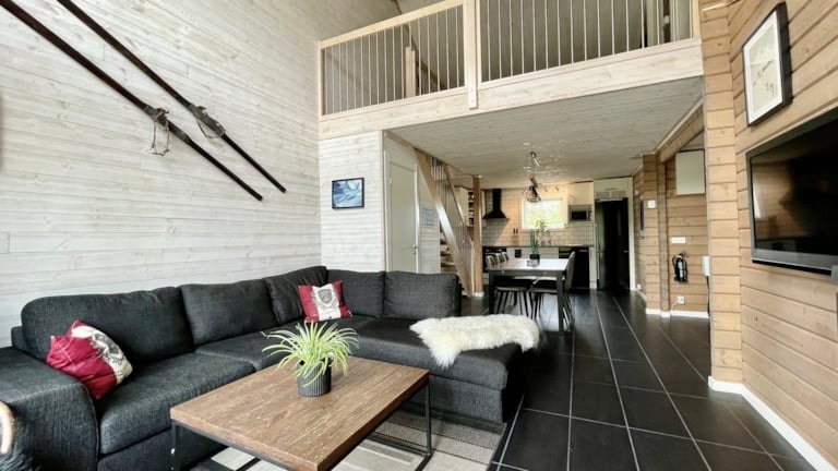 Moderne stue med lyse trævægge, sort sofa, sofabord og tæppe i fåreskind. Ski er monteret på væggen, og der er en hems over et spisebord og køkken i baggrunden.