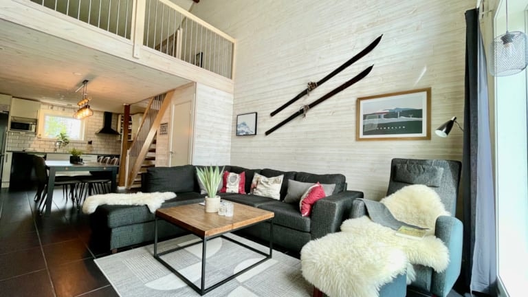 Moderne stue med hems, grå sofa, sofabord i træ, pelsdække, vægmonterede vintageski, stort vindue og åbent køkken i baggrunden. Lyse trævægge og sorte detaljer skaber et hyggeligt og stilfuldt rum.