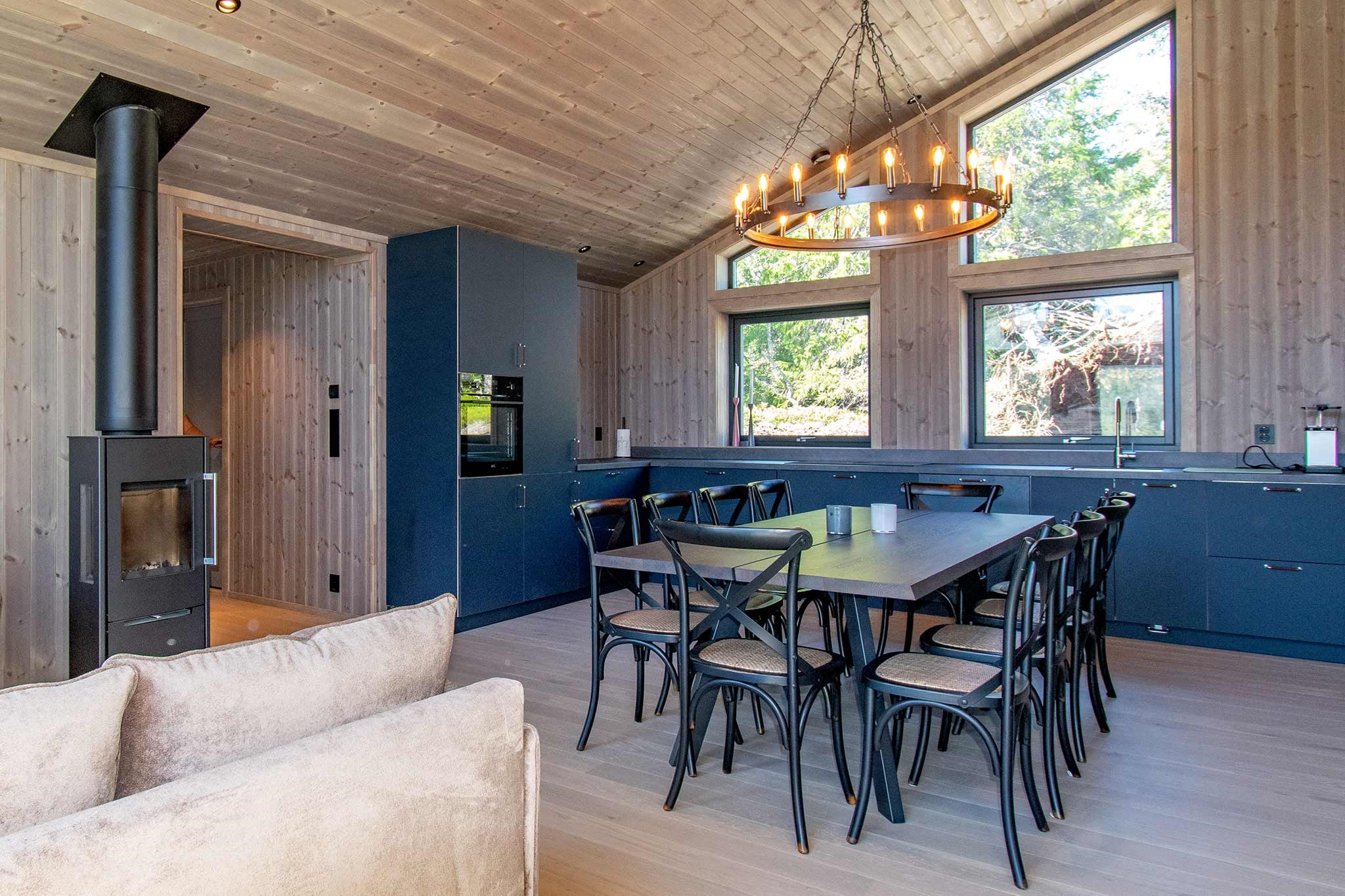 Moderne åbent køkken og spisestue med mørkeblå skabe, sort spisebord og stole, store vinduer, træpaneler på vægge og i loftet, en beige sofa, pejs og en cirkulær lysekrone.