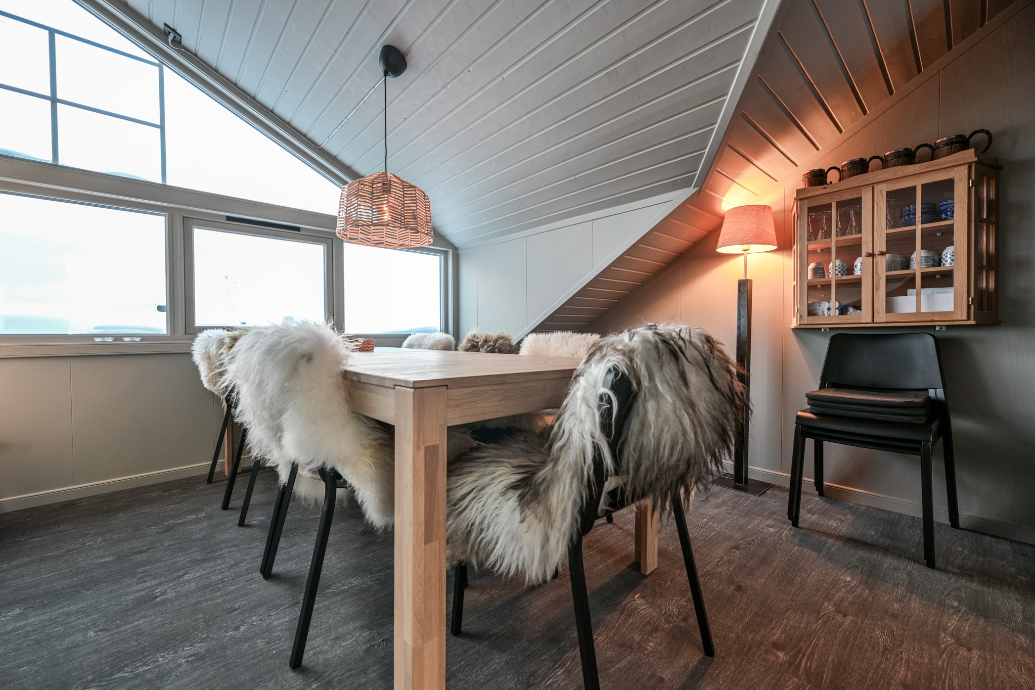 En hyggelig spisestue med et træbord, sorte stole beklædt med pelstæpper, en pendellampe ovenover, en gulvlampe, et væghængt skab med krus og skrå lofter med hvide paneler.