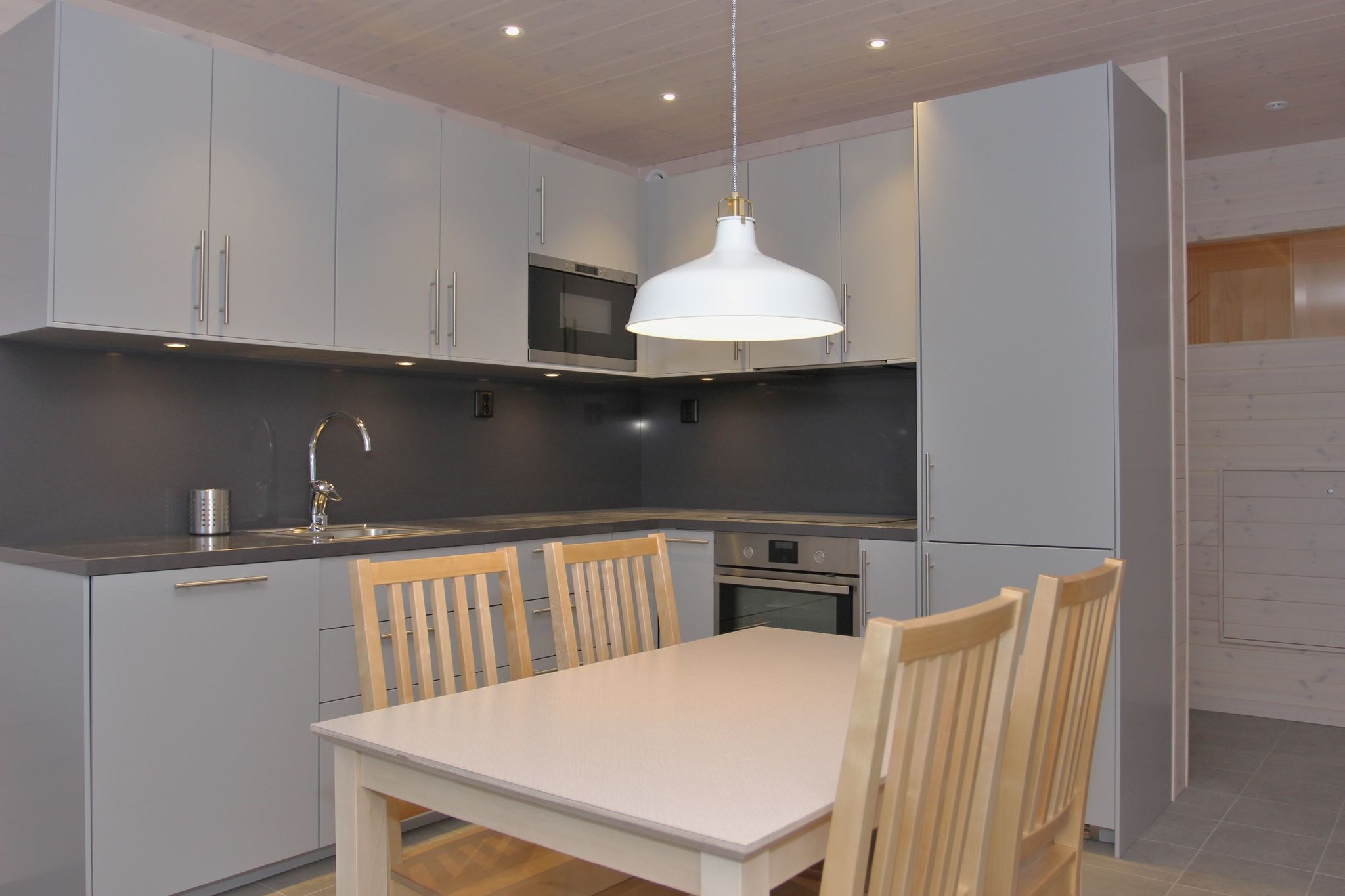 Et moderne køkken med lysegrå skabe, indbygget ovn, mikrobølgeovn og vask. Et spisebord i træ med fire matchende stole står under en hvid hængelampe. Rummet har et minimalistisk og rent design.