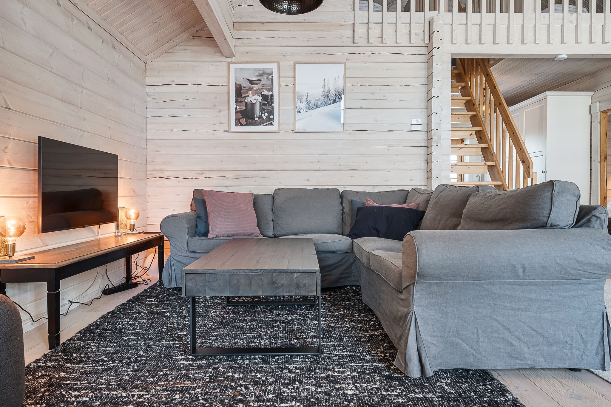 En hyggelig stue med en grå L-formet sofa, sort tæppe, sofabord i træ, væghængt TV og varm belysning. Lyse træpaneler på vægge og i loftet, to indrammede fotos og en trappe, der fører til en øvre etage, er synlige.