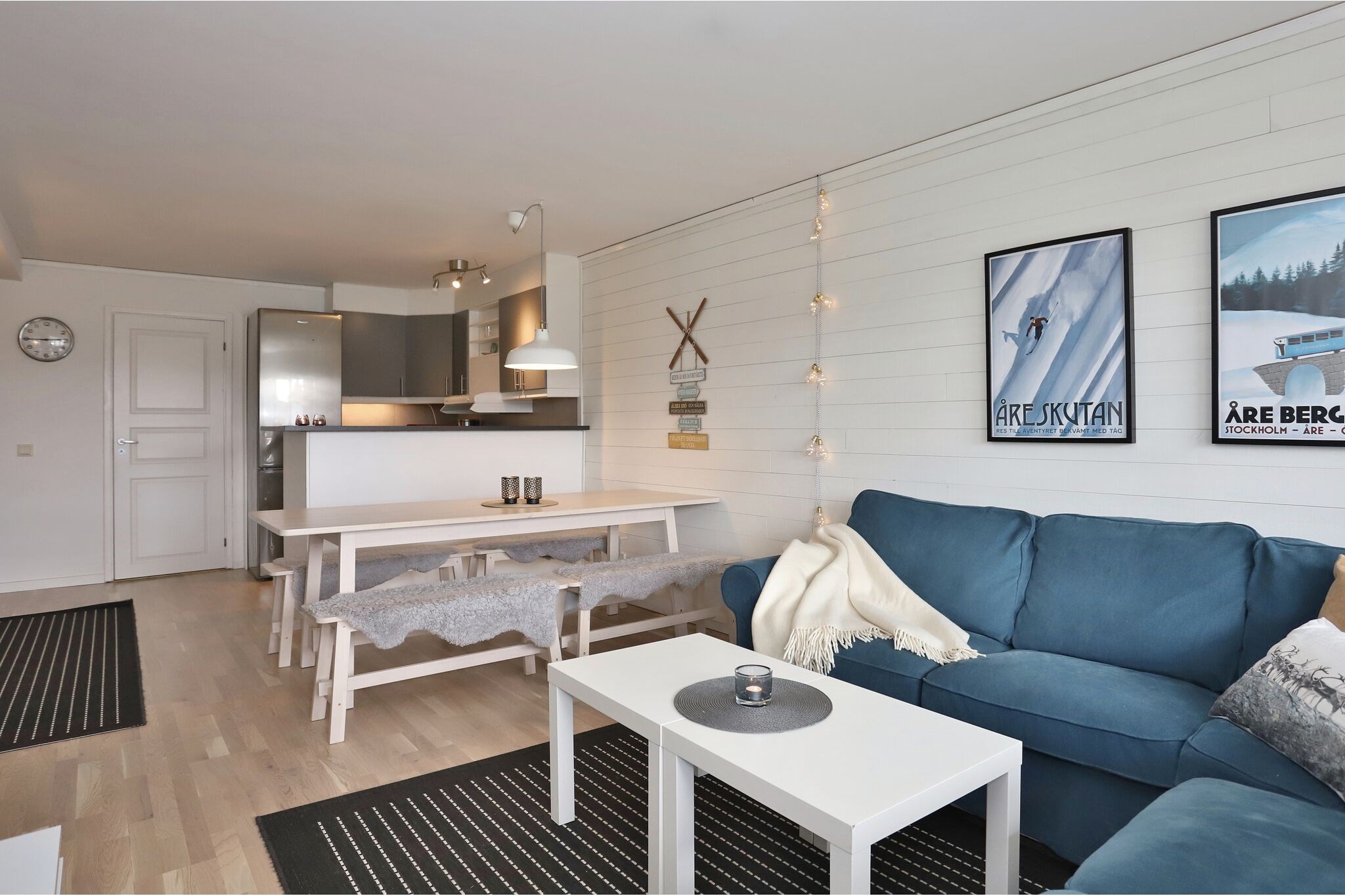 En moderne stue og spisestue med en blå sofa, et hvidt sofabord, et spisebord med bænke beklædt med pelsdække, vægkunst og et køkken i baggrunden. Lyst trægulv og neutral indretning skaber en hyggelig atmosfære.