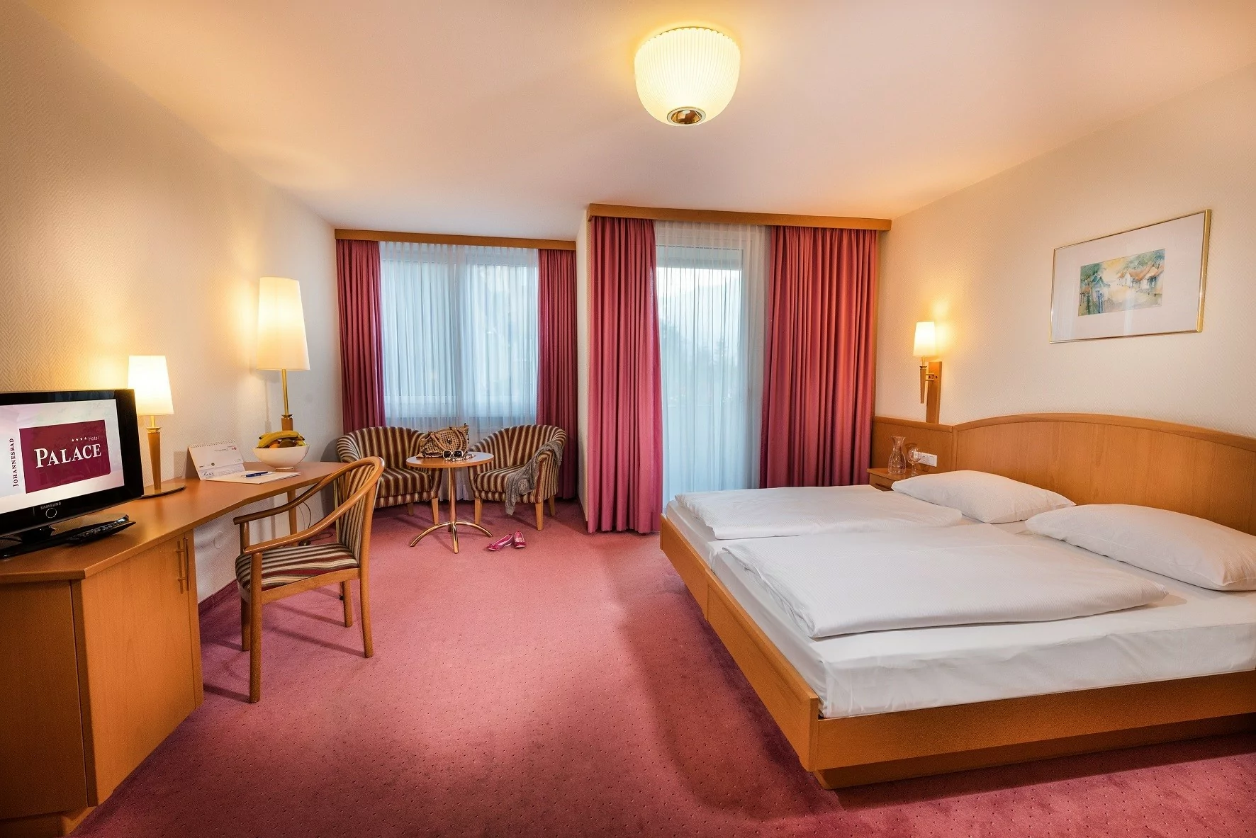 Et hotelværelse med en dobbeltseng, to zebrastribede stole og et lille bord ved vinduet, et skrivebord med et tv og lyserødt tæppe og gardiner, der skaber en hyggelig og varmt oplyst atmosfære.