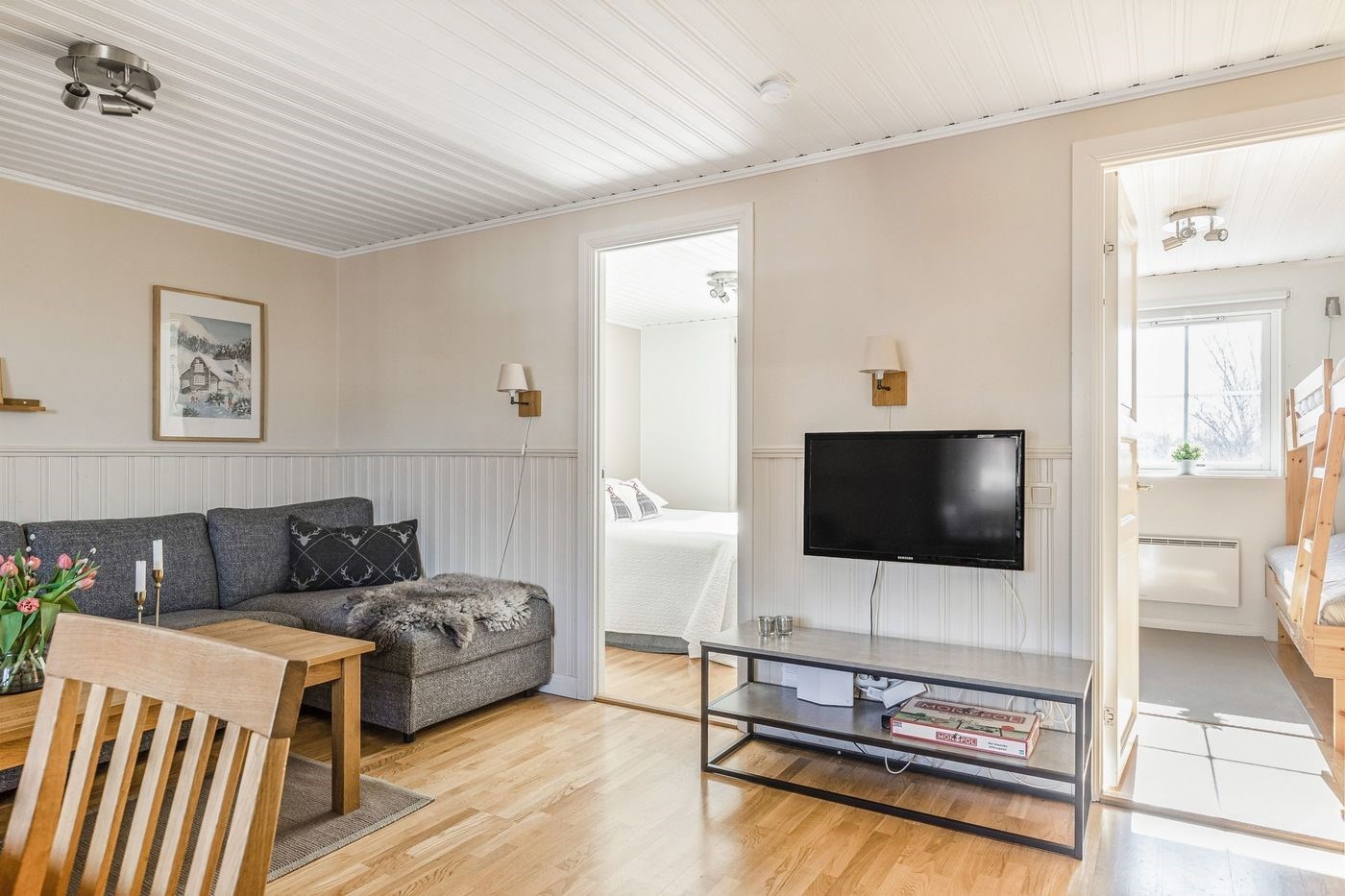 En hyggelig stue med en grå sofa, et spisebord i træ, et tv på et glasbord og lyse trægulve. To døråbninger fører til et lyst soveværelse og et værelse med køjesenge. Sollys strømmer ind gennem vinduerne.