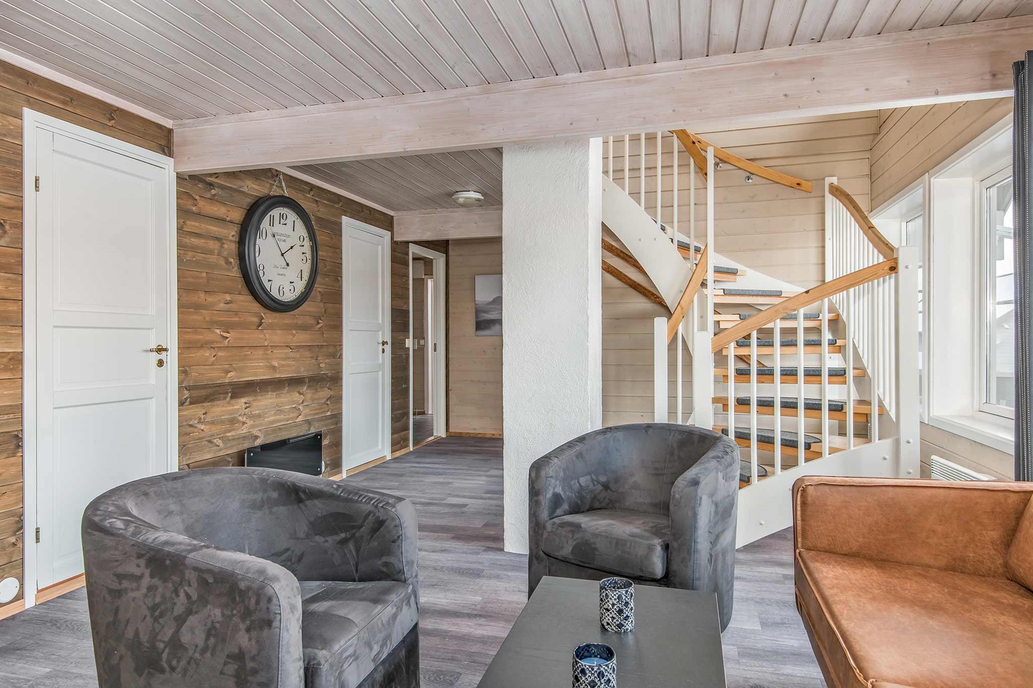 Moderne stue med træpaneler på væggene, en vindeltrappe, to grå fløjlsstole, en brun læderlænestol, et sort sofabord med stearinlys, et stort vægur og store vinduer, der lukker naturligt lys ind.