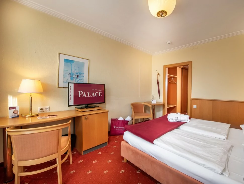 Et hyggeligt hotelværelse med en enkeltseng, rødt tæppe, træmøbler, et skrivebord med en lampe, et tv med teksten "PALACE", en stol, et skab og en badekåbe på sengen. Værelset fremstår ryddeligt og indbydende.